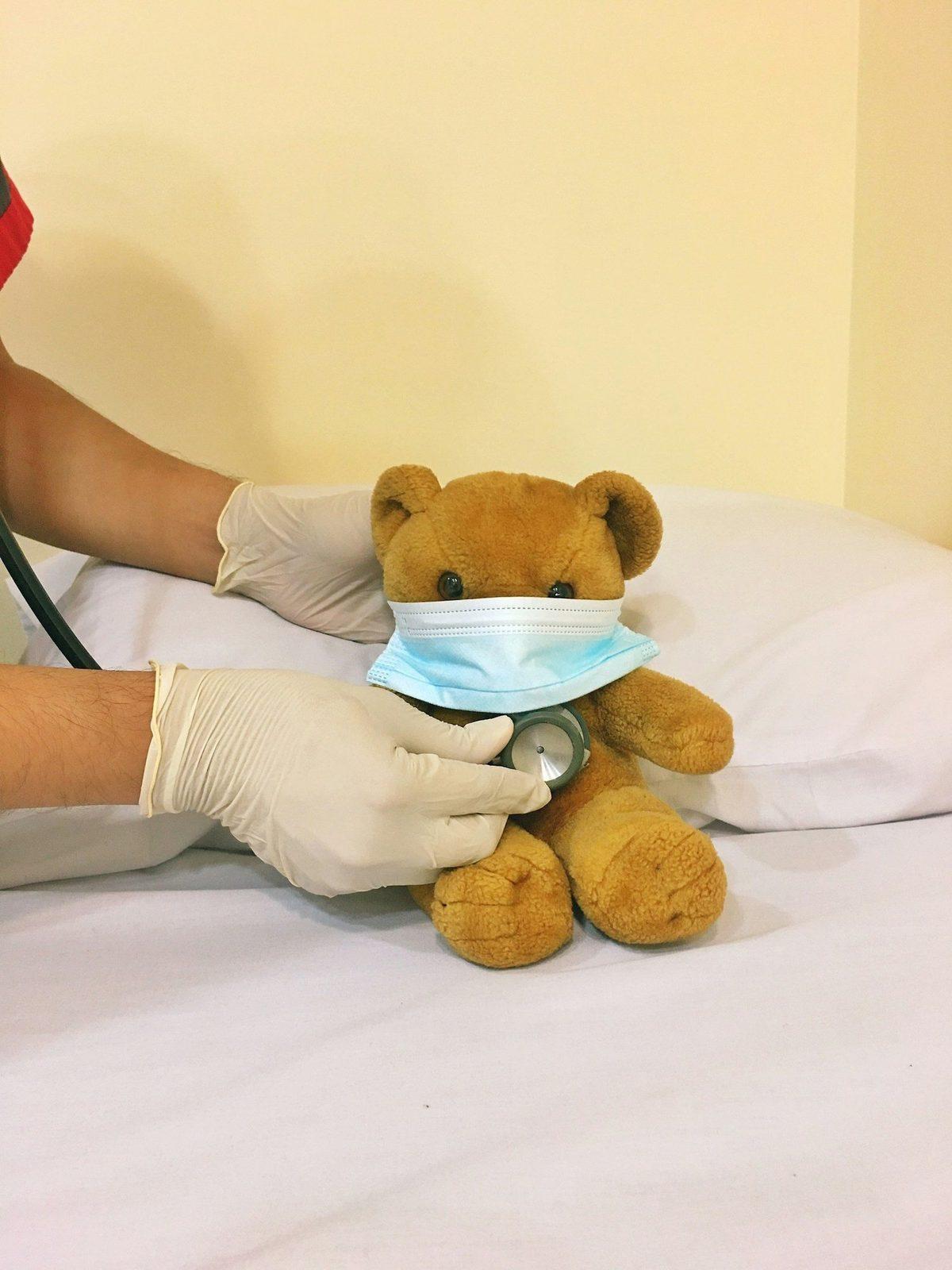 A gloved hand placing a face mask on a teddy bear
